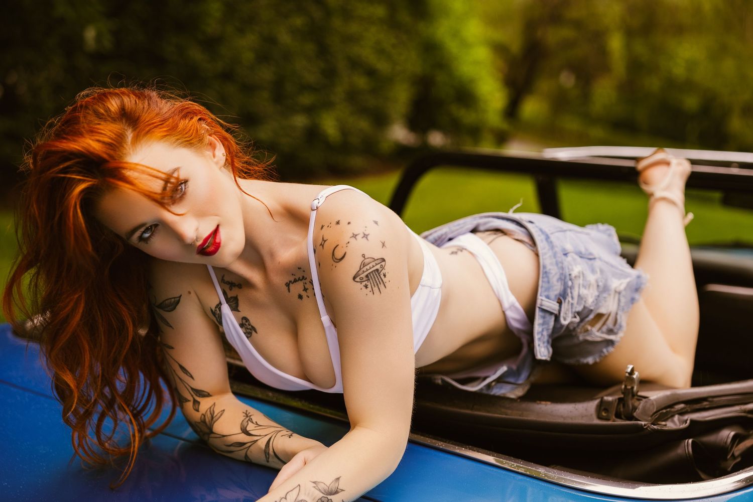 Tattooed redhead woman posing on vintage convertible car hood outdoors in summer setting.
