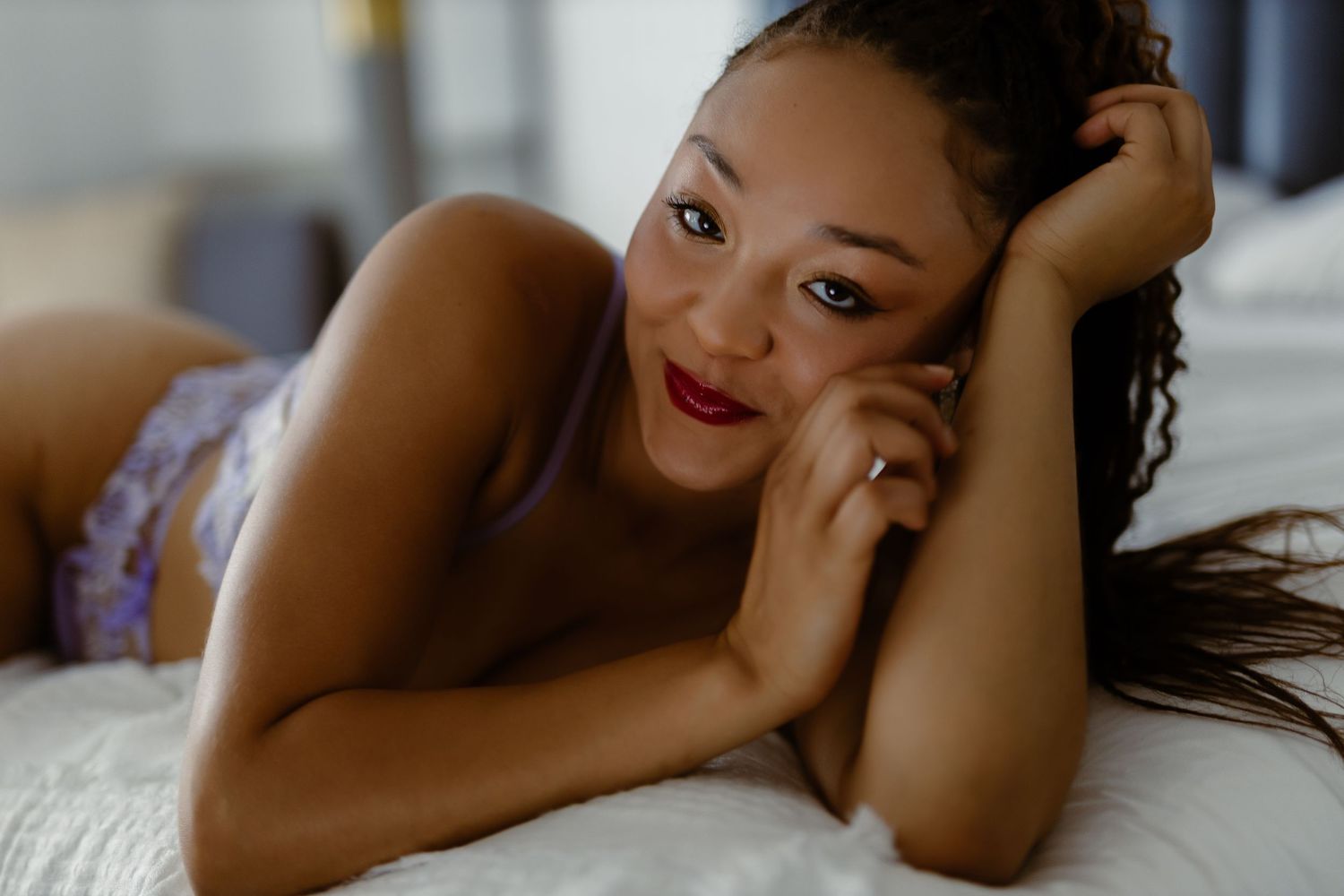 A person with red lipstick lying on white bedding in a relaxed boudoir pose.