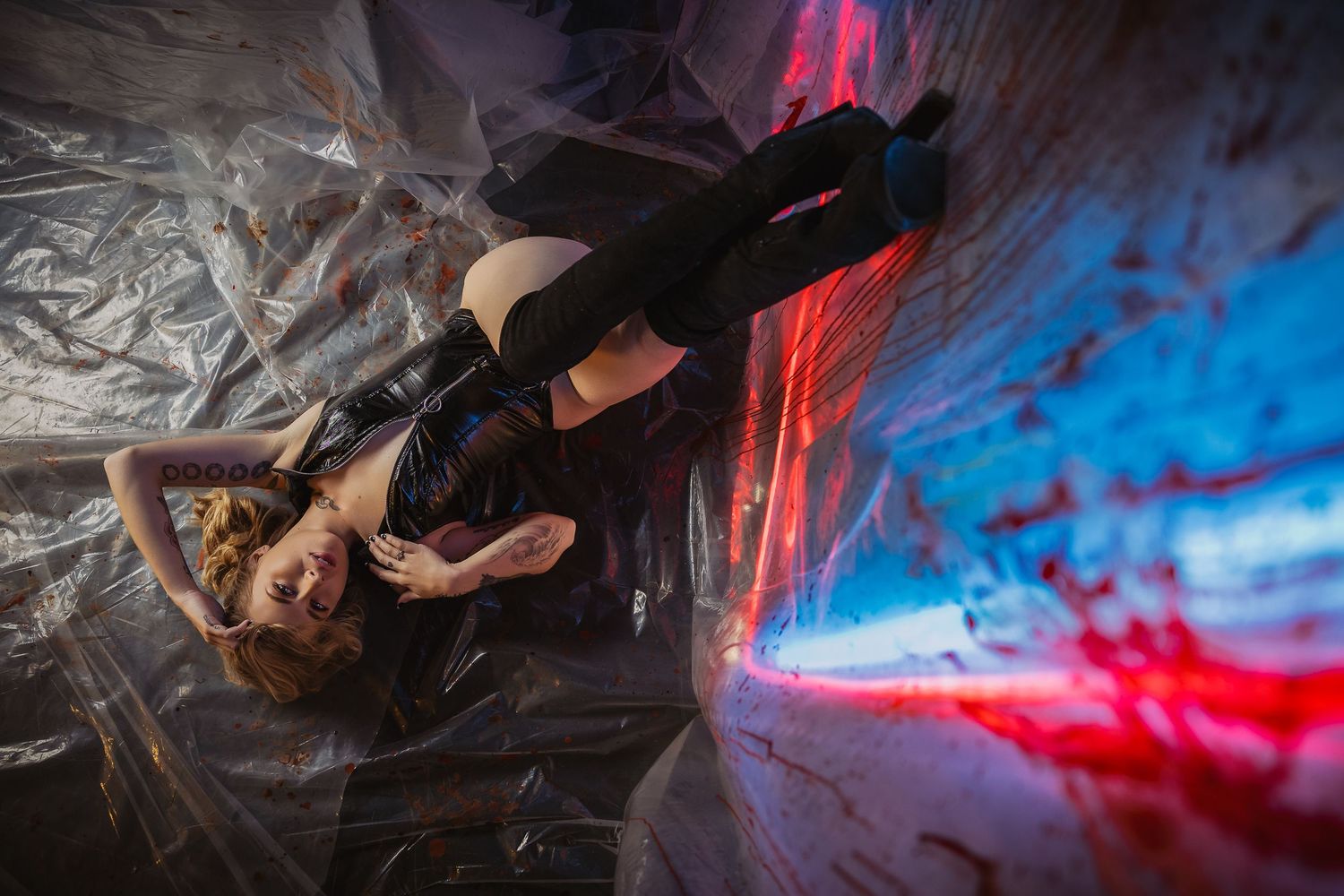 Woman in black outfit lying on plastic sheeting with dramatic red and blue neon lighting in artistic photo shoot.