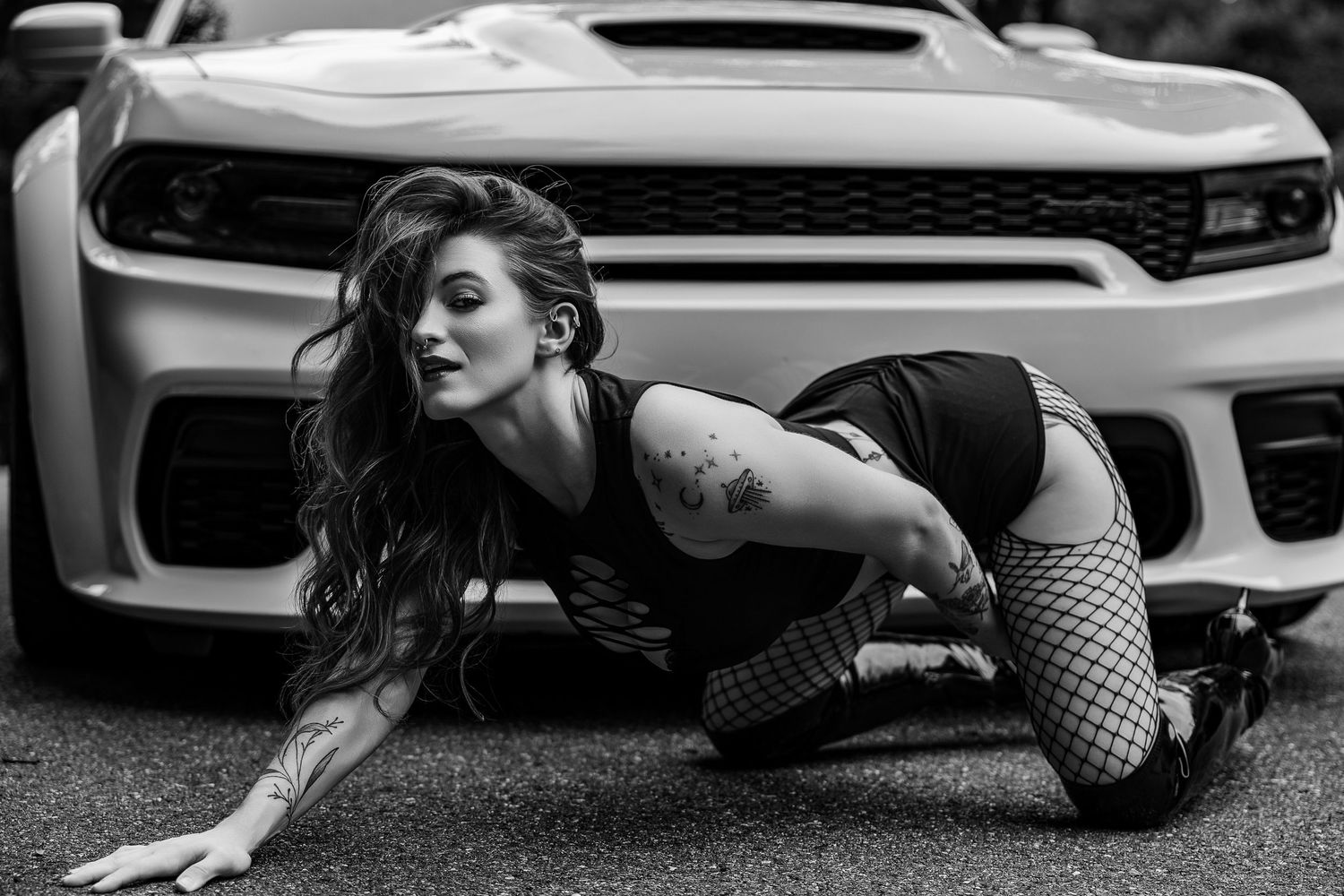 Black and white photo of a tattooed woman in fishnet stockings posing in front of a Chevrolet Camaro.