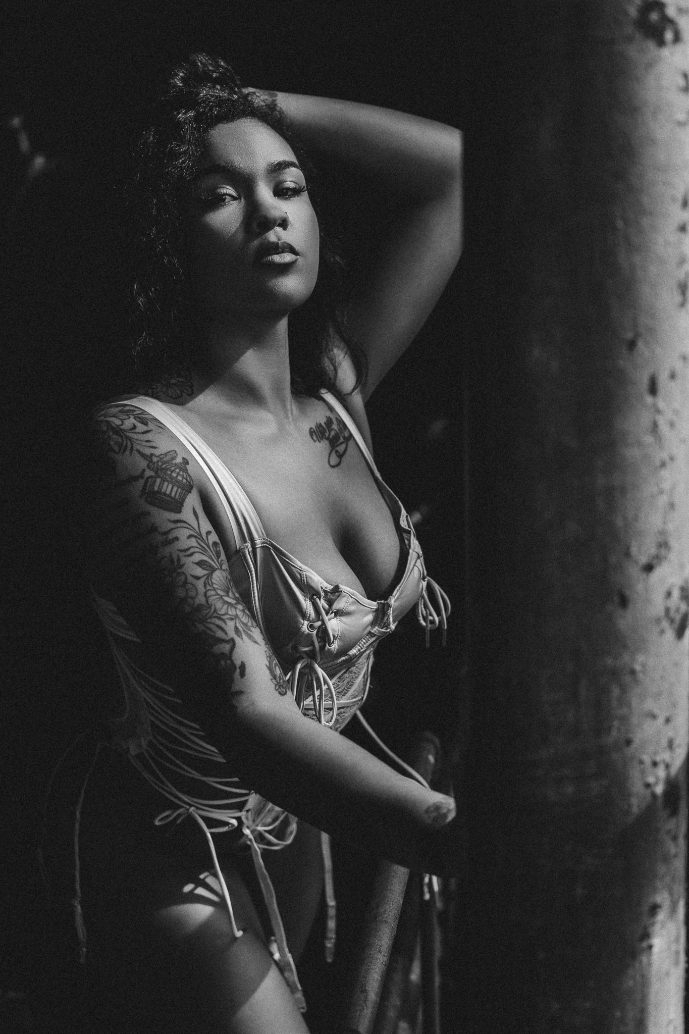 Black and white portrait of a tattooed woman in lingerie posing dramatically in moody low-key lighting.