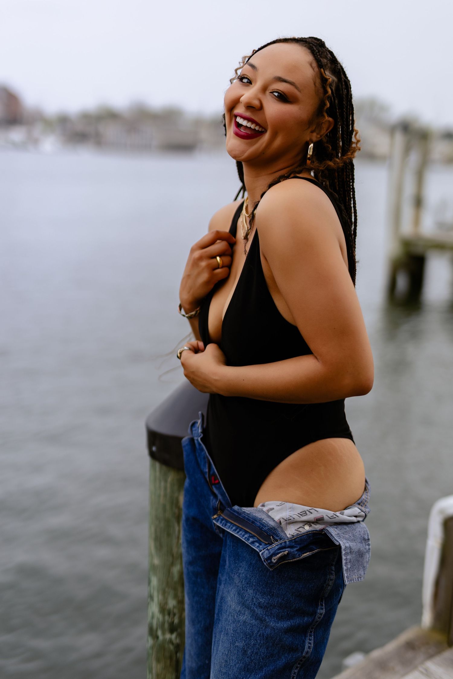 Joyful person wearing a black swimsuit and jeans poses by a dock on a cloudy day near the water in Annapolis, outdoor boudoir.