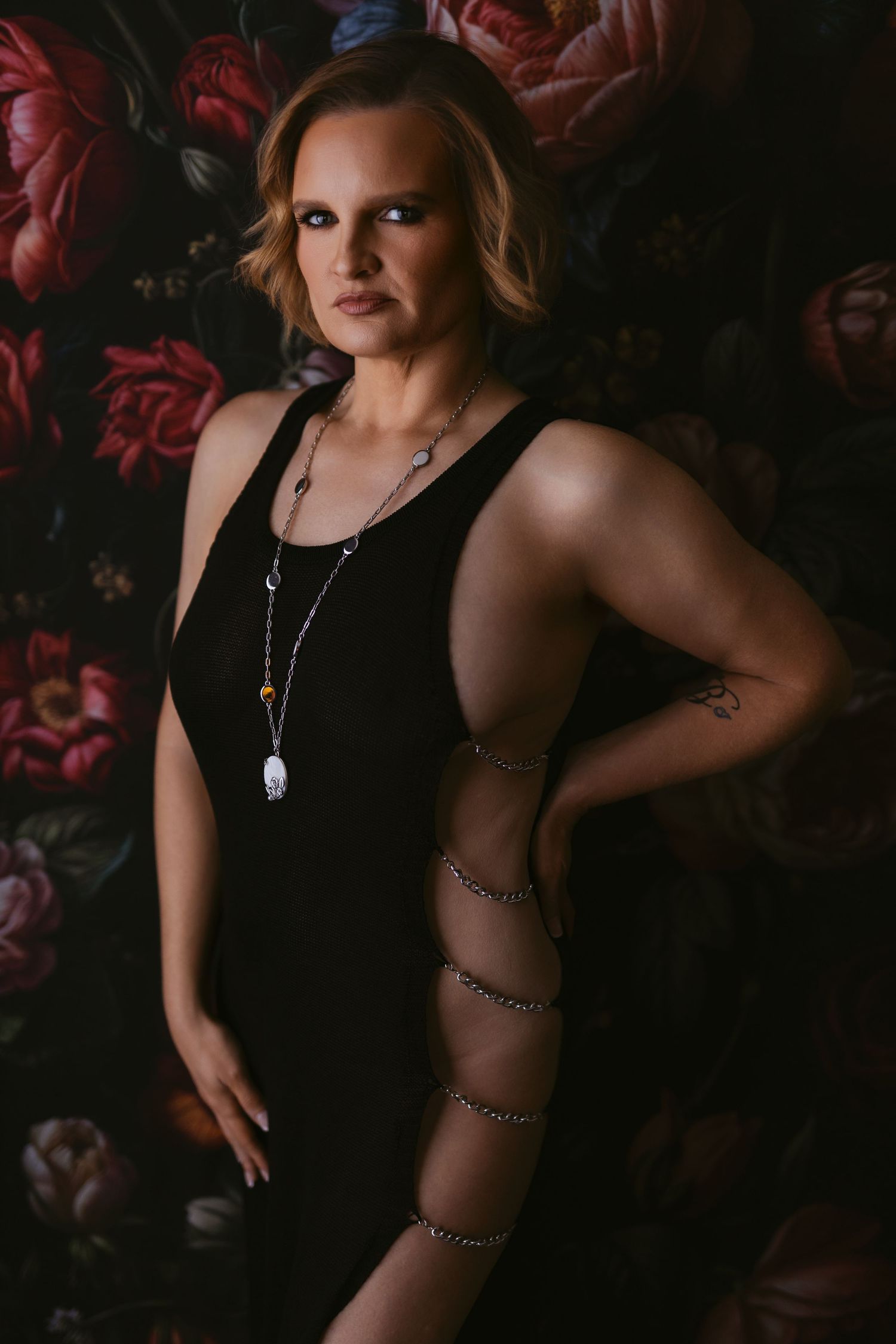 A moody boudoir photoshoot features a figure in black against a dark floral backdrop with dramatic lighting.
