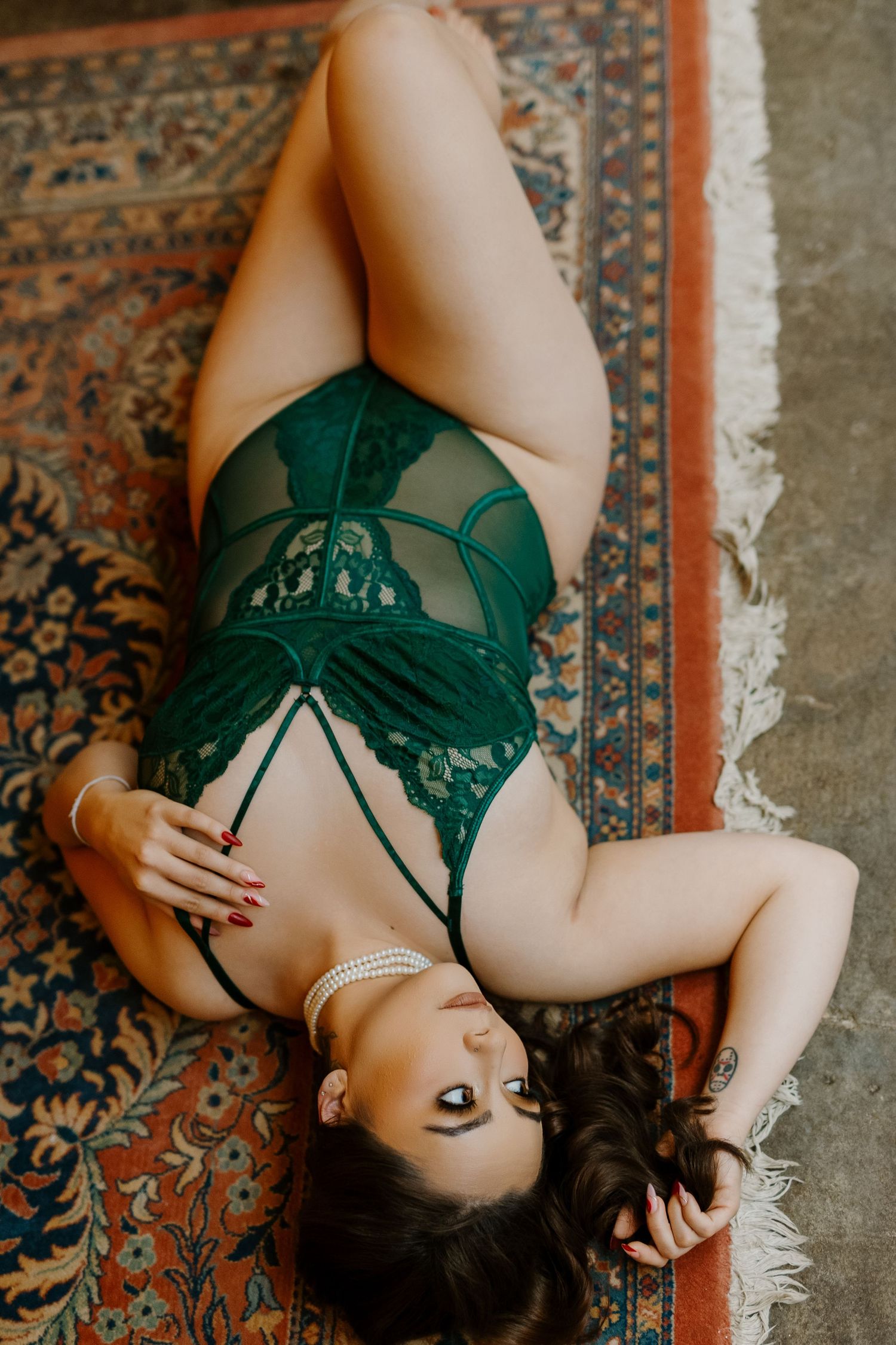 Woman in green lace lingerie bodysuit posing on a vintage patterned rug in a boudoir photography session.