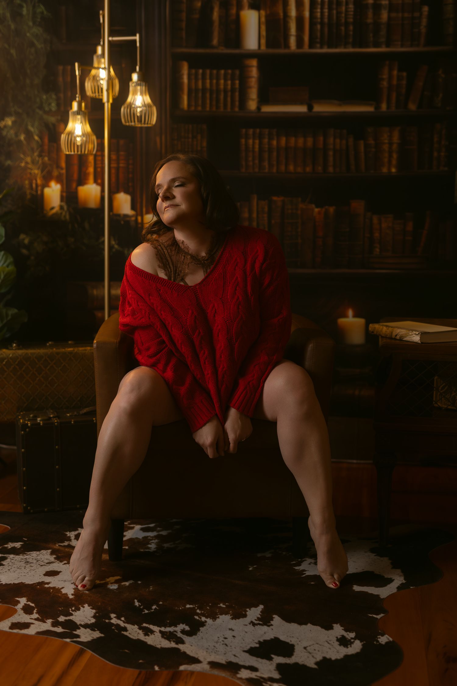 Moody boudoir scene in library with red sweater and hanging Edison bulbs casting warm glow against bookshelves.