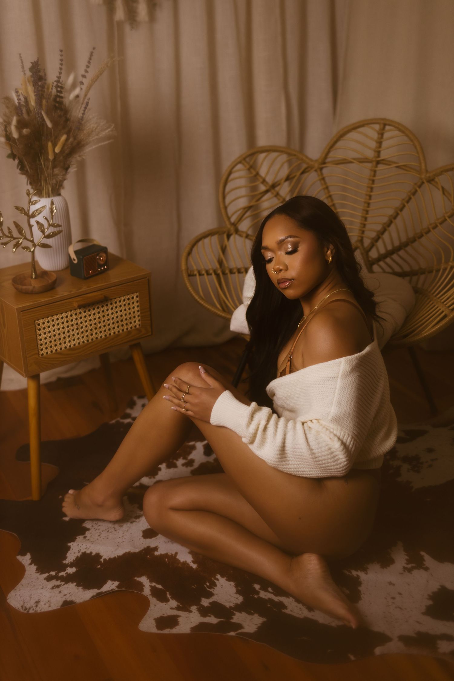 Moody boudoir photo with rattan peacock chair, side table with dried flowers, and cozy white sweater in warm sepia tones.