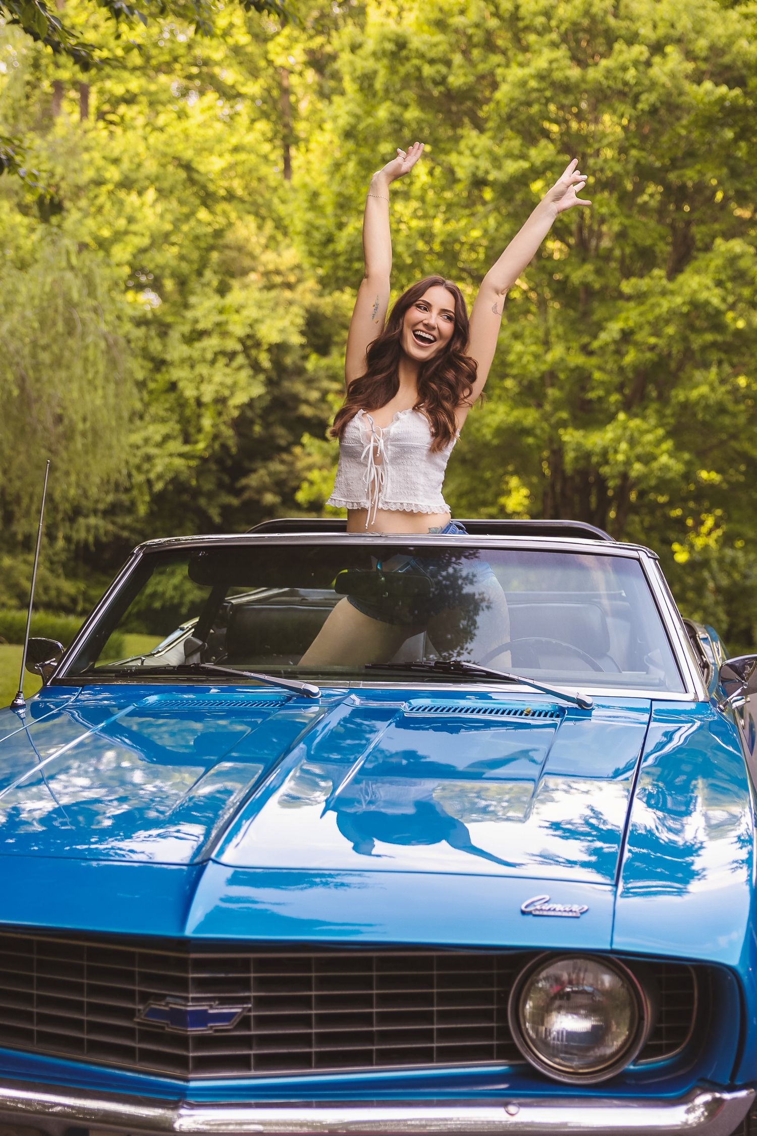 Joyful woman with arms raised standing in blue classic Camaro convertible surrounded by lush green trees.