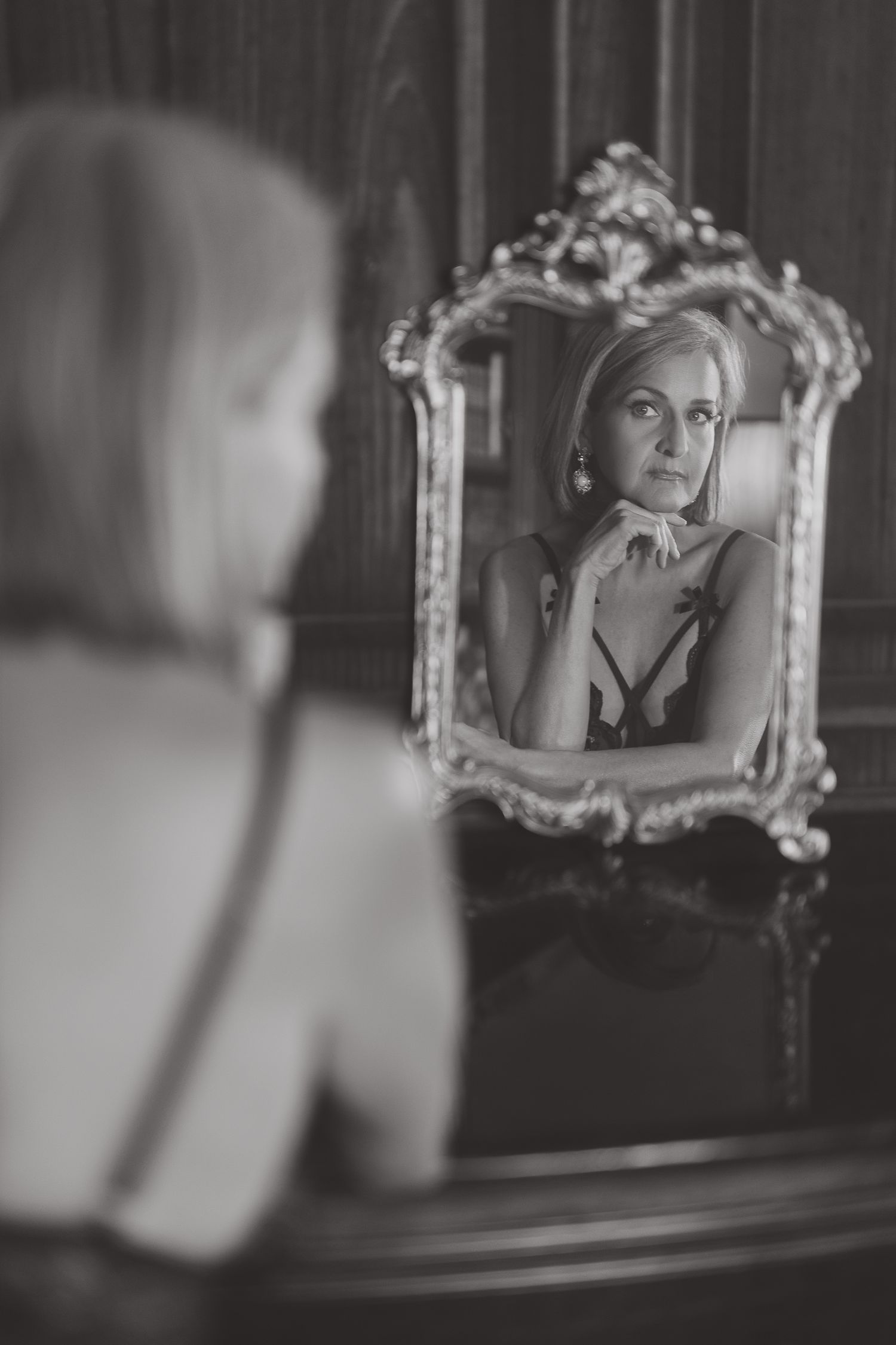Black and white portrait of a woman in lingerie gazing at her reflection in an ornate vintage gold-framed mirror.