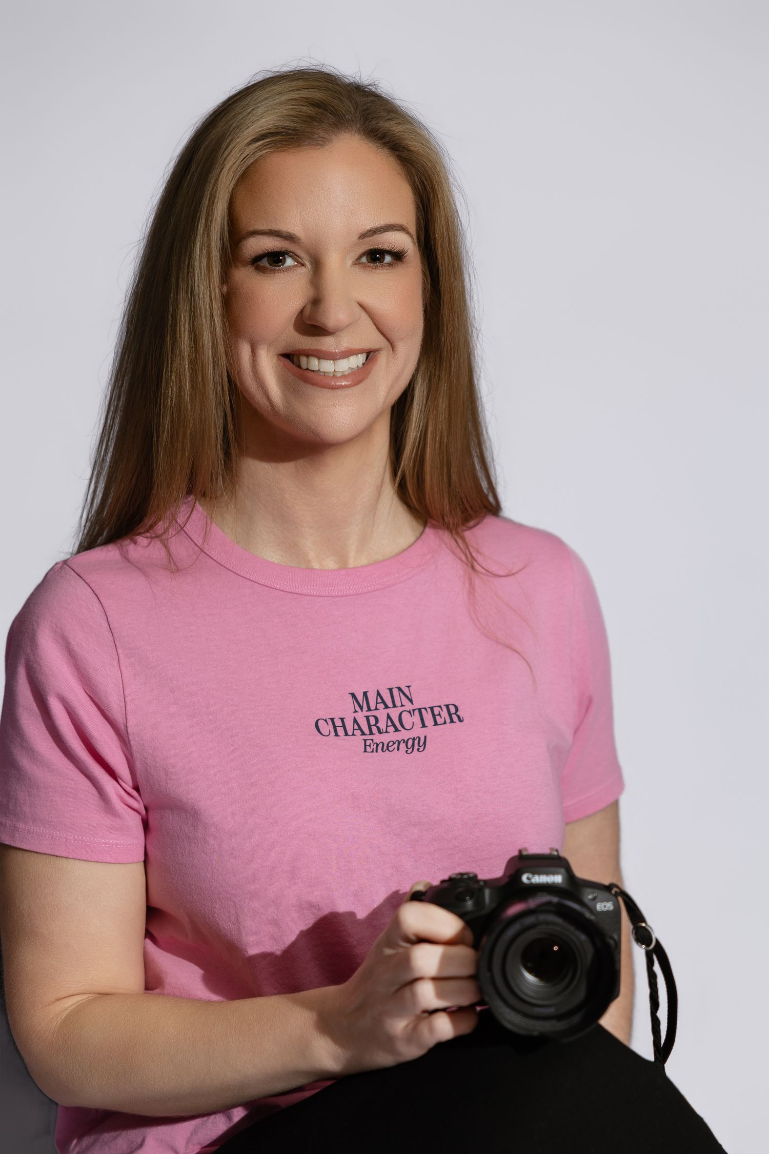 Smiling woman in pink Main Character Energy t-shirt holding a Canon camera against white background.