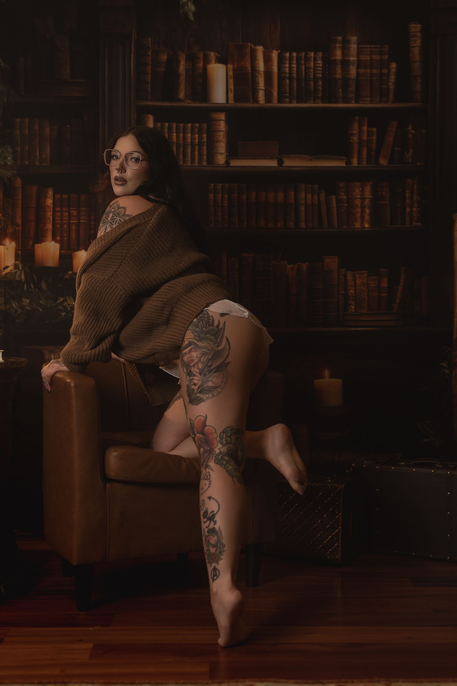 Tattooed woman in brown sweater posing in a dark library with bookshelves and candles in the background.