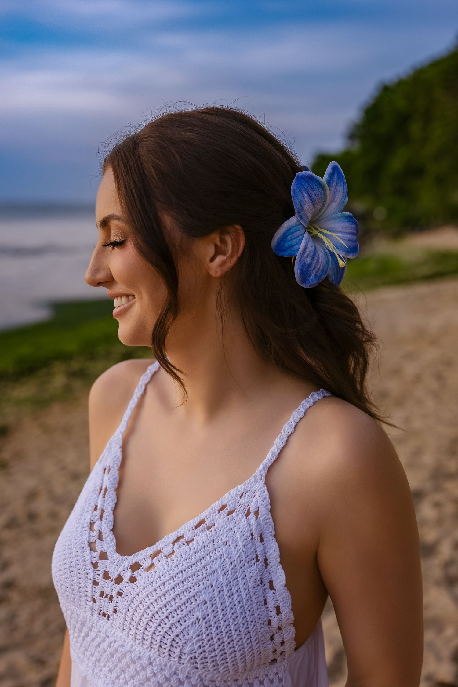 Smiling brunette woman wearing white crochet top with blue flower hair accessory on a tropical beach.