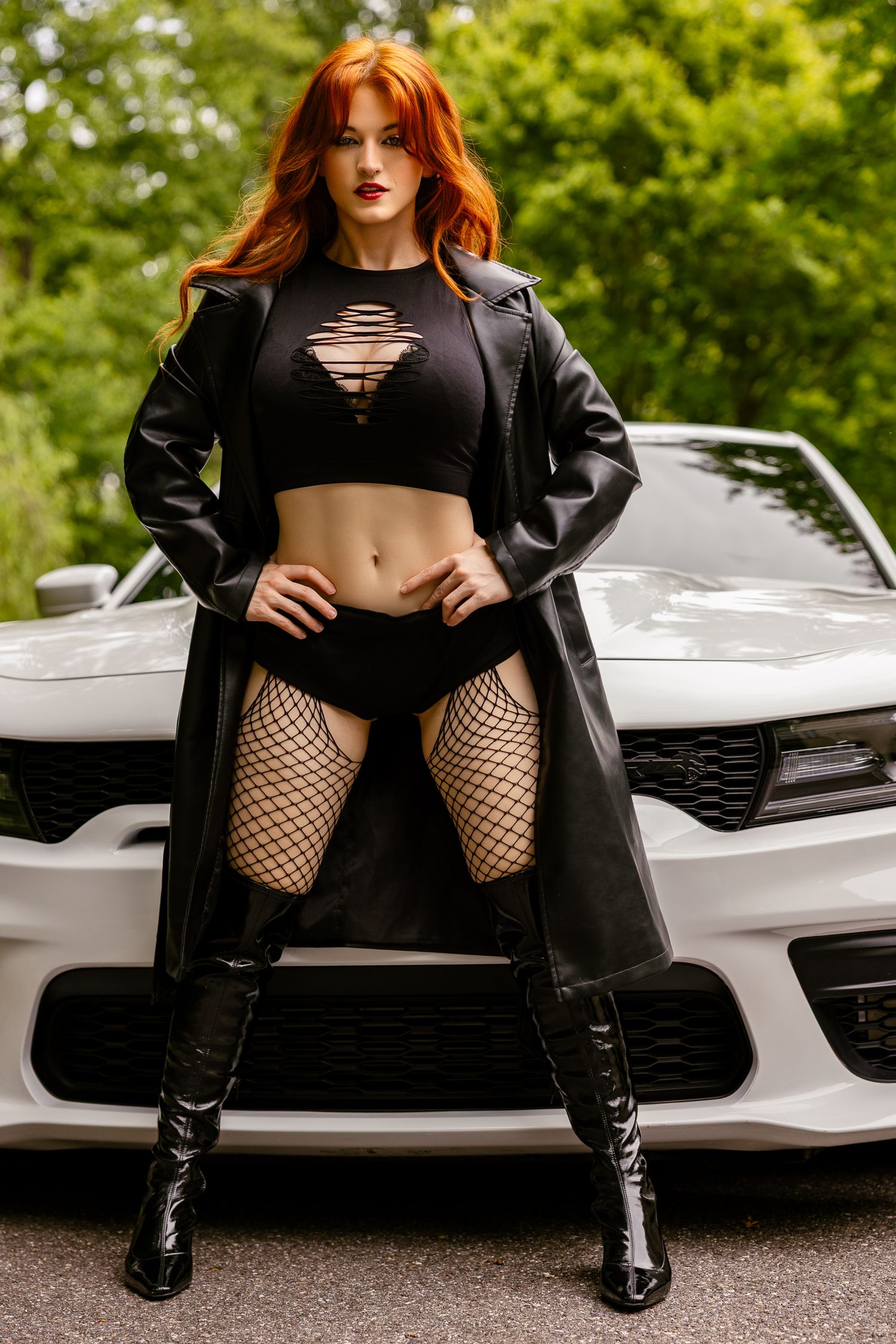 Red-haired woman in black crop top, fishnet tights, leather coat and boots posing in front of a white sports car.