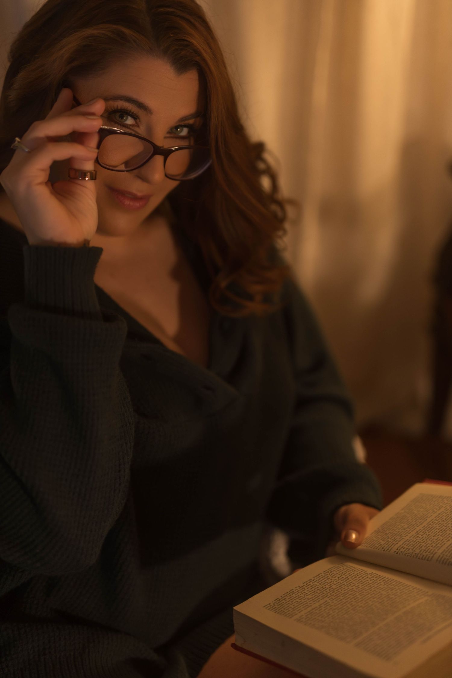 A cozy reading scene with someone wearing glasses and a dark sweater browsing through a book in warm lighting.