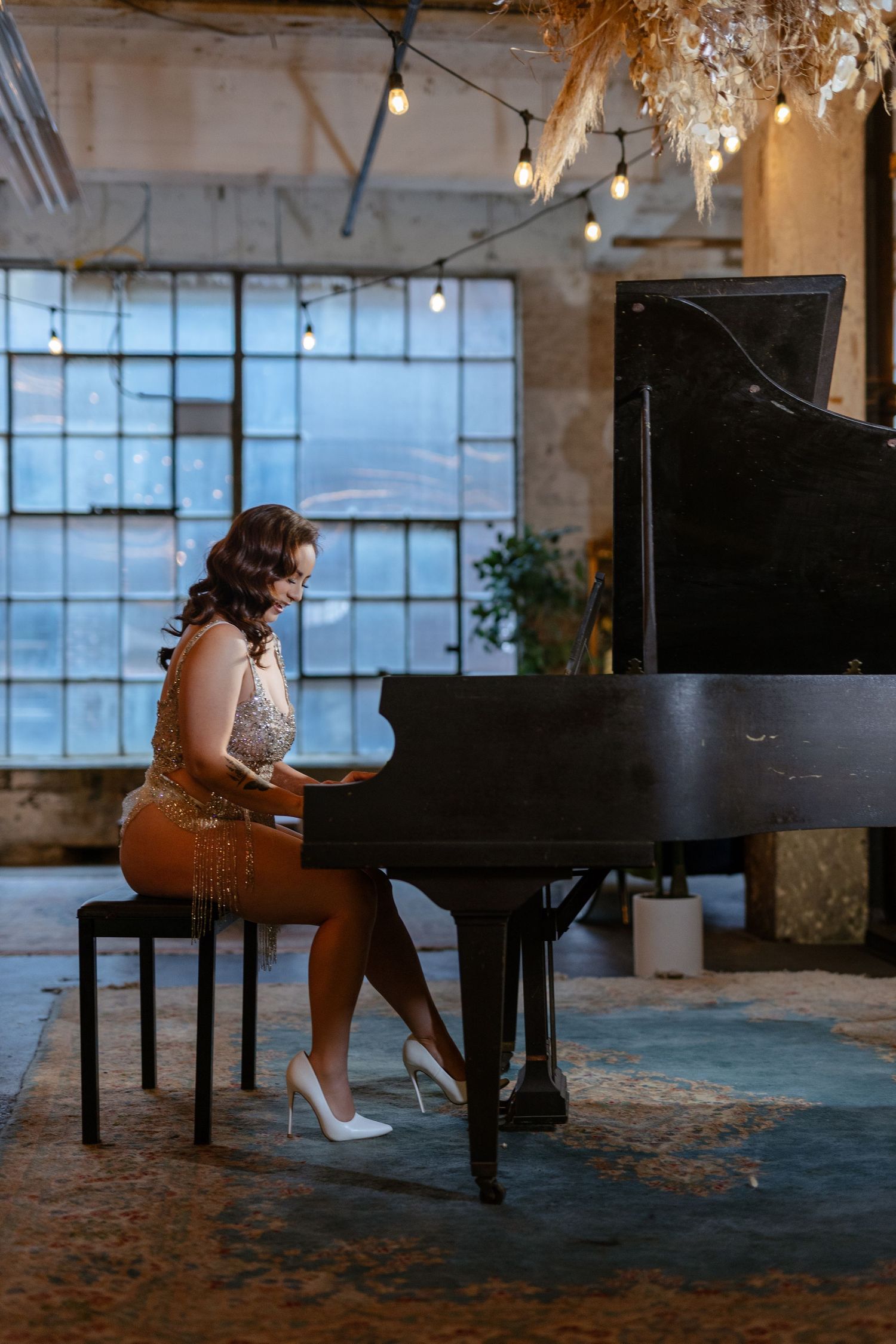 Elegant woman in gold dress playing a black grand piano in a stylish loft with string lights and large windows.