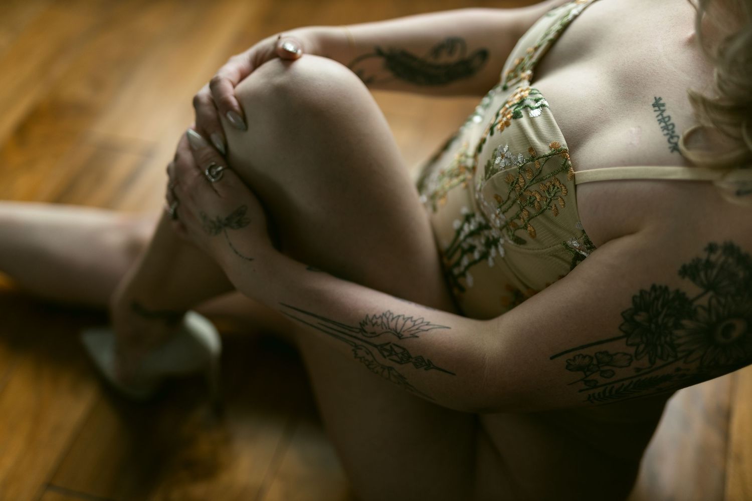 Intimate boudoir photo showing delicate lace lingerie details against hardwood flooring, woman with warrior tattoo over breast cancer scar.
