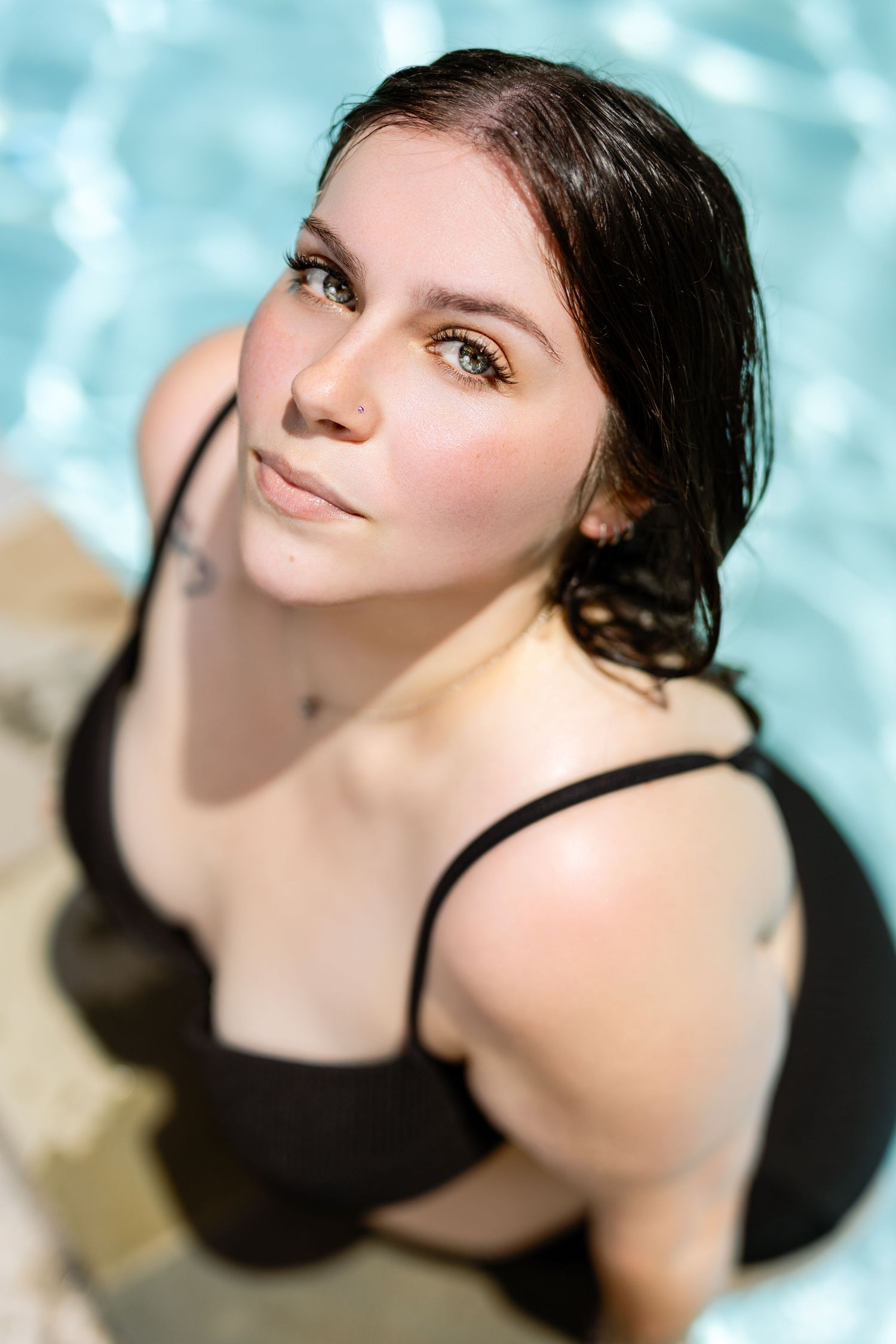 Young woman with wet dark hair in black bikini top poses by pool edge in bright sunlight.