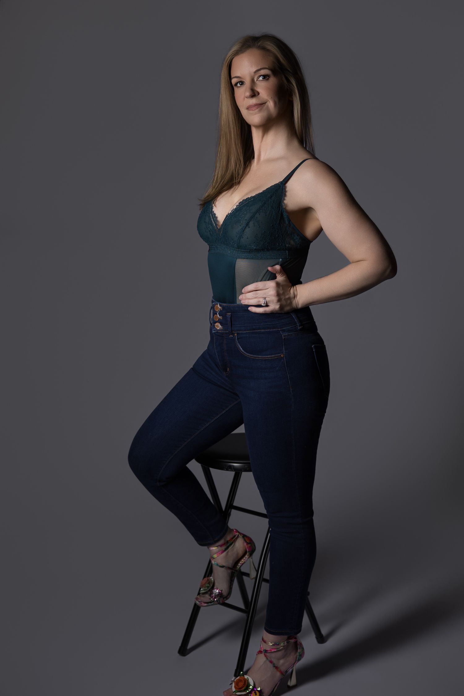 Woman in teal bodysuit and dark jeans poses confidently on a stool against a gray studio background.