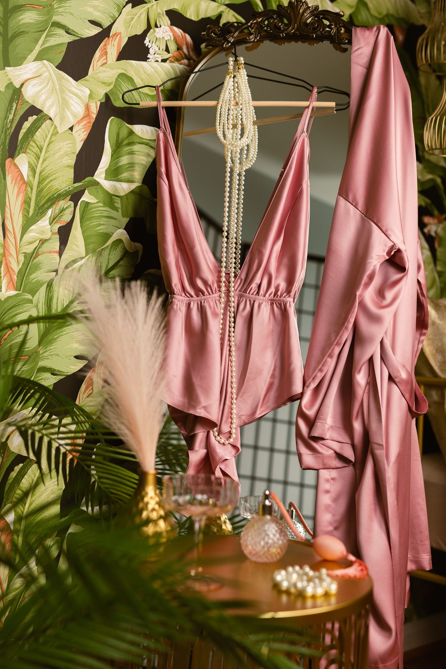 Pink silk robe and lingerie hanging on a hanger surrounded by tropical leaves and decorative elements against a dark background.