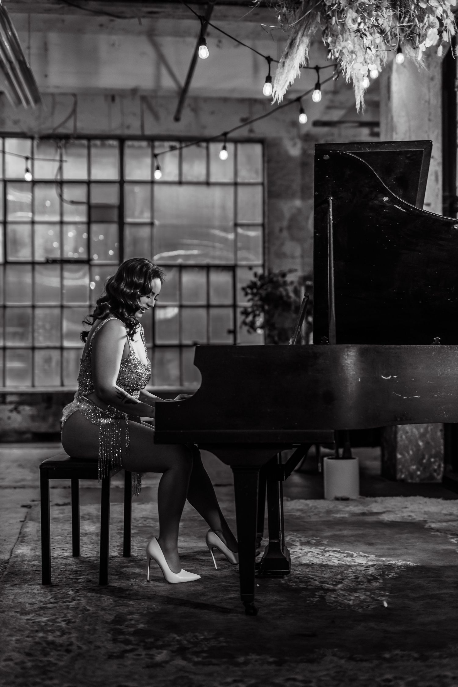 A pianist in a sparkly dress performs at a grand piano in a moody industrial warehouse setting with large windows.