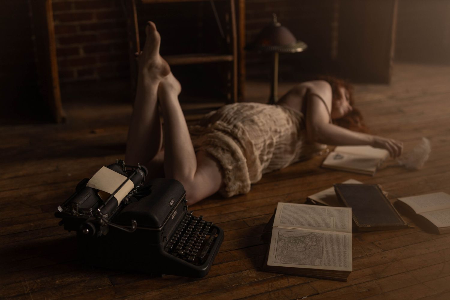 A person lying on hardwood floor next to a vintage typewriter and papers in moody lighting.