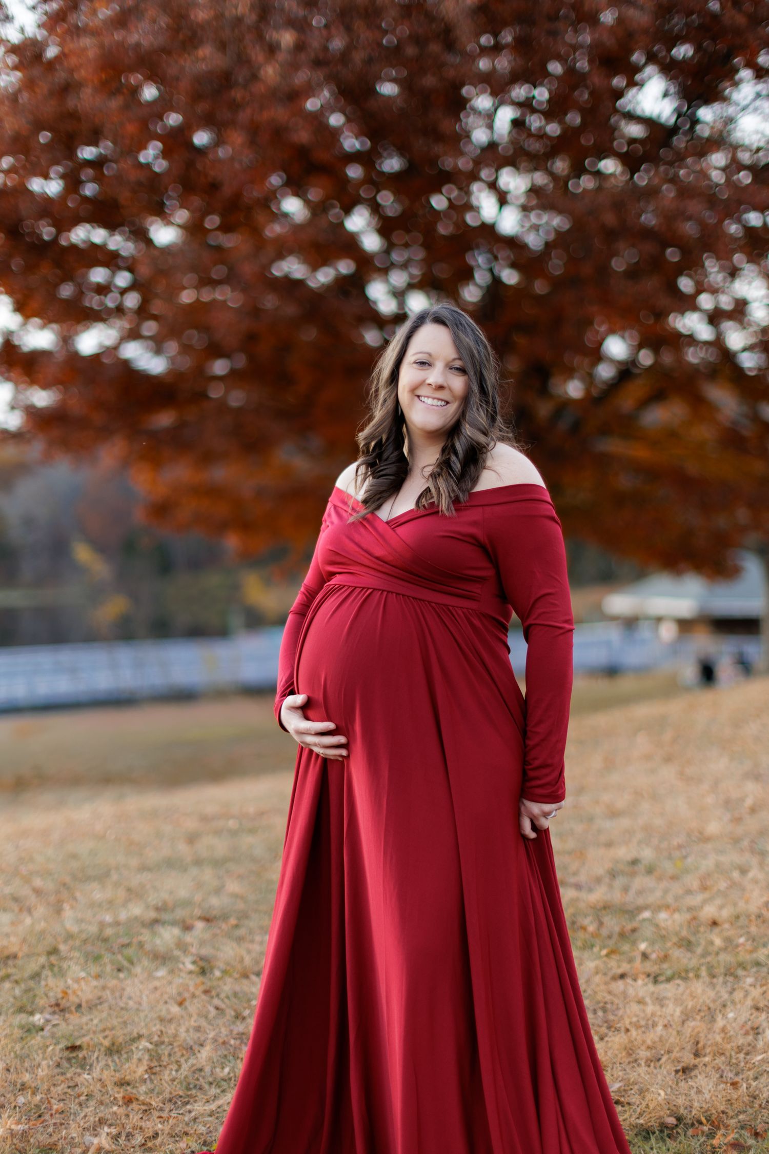 A pregnant woman in a deep red dress stands in a field of closely cropped grass with fall foliage behind her.