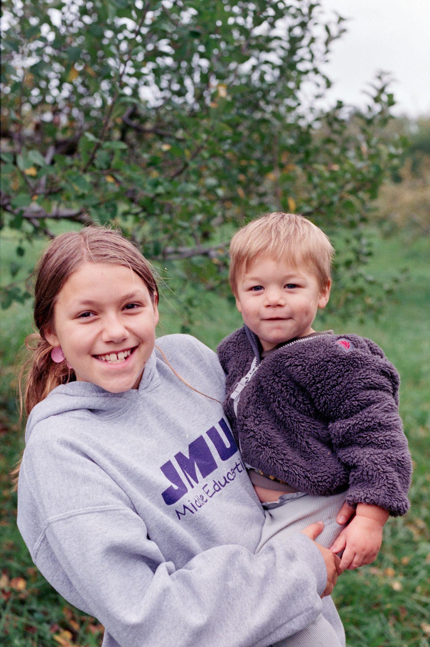 A girl in an apple orchard holds her brother as they smile at the camera.