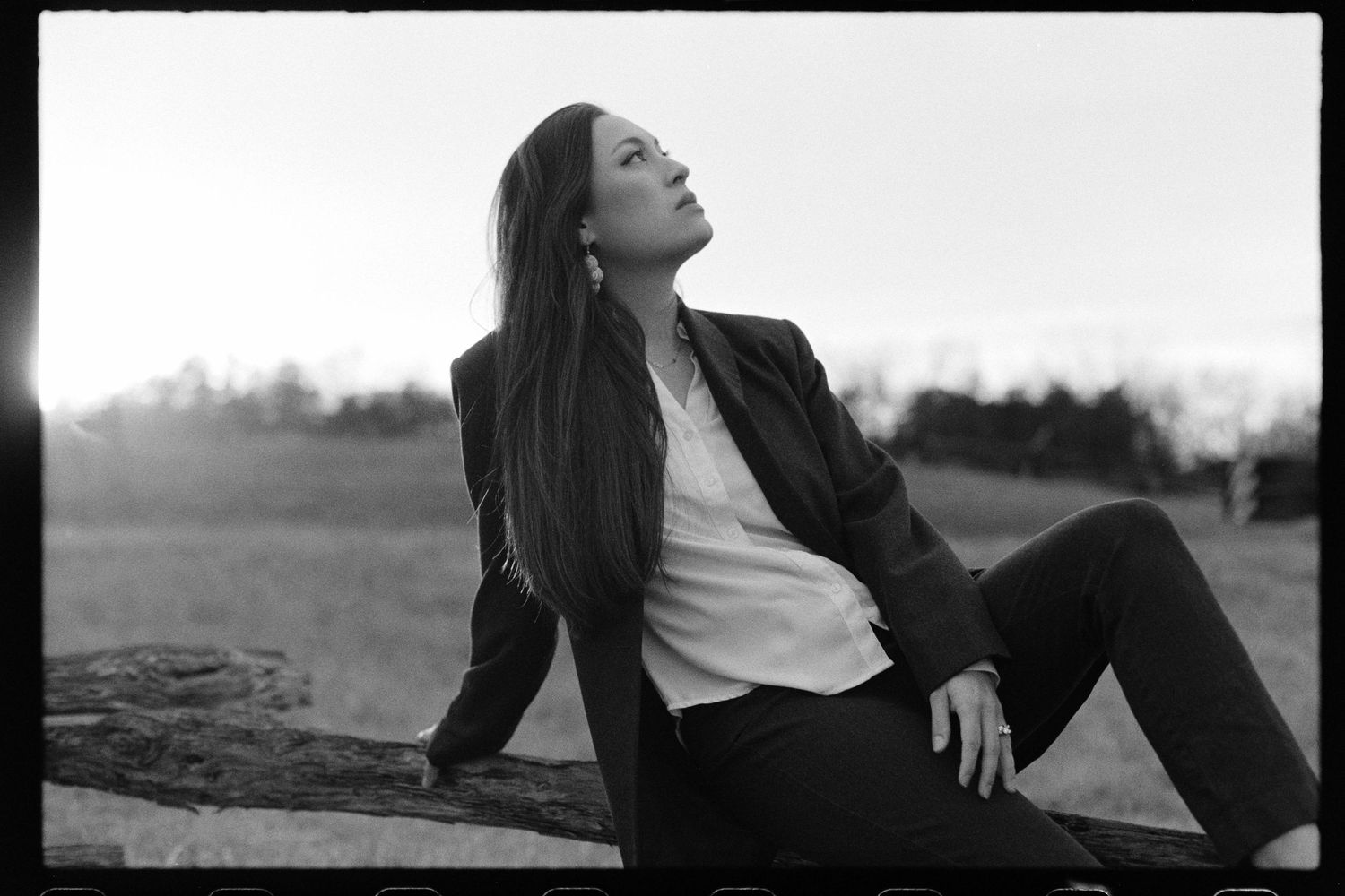 Black & white portrait of a woman reclining on a wooden fence in a field. She's looking up at the sky.