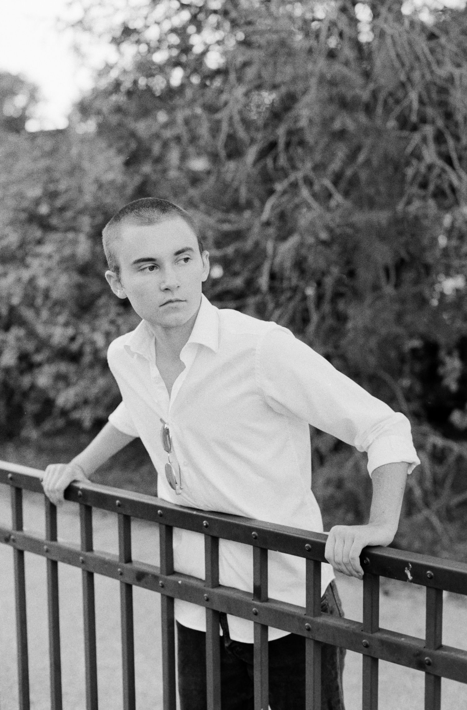 Black & white portrait of a young man leaning on a metal chest-high fence supporting himself with his arms.