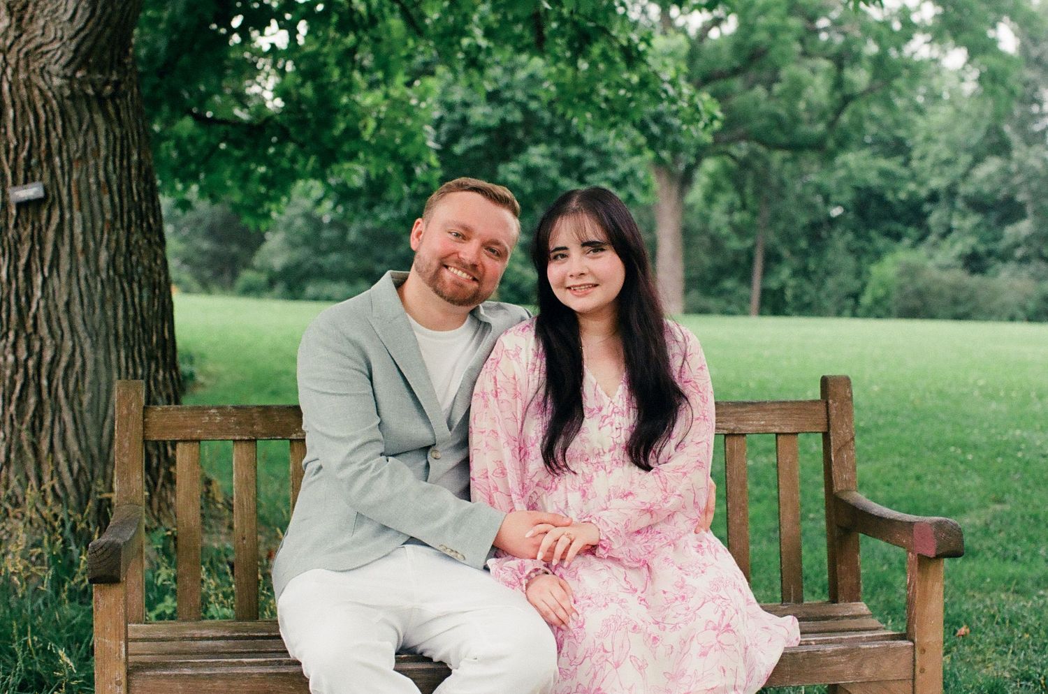 A man and a woman sitting on a wooden bench in a park. They are holding hands to show off her engagement ring.