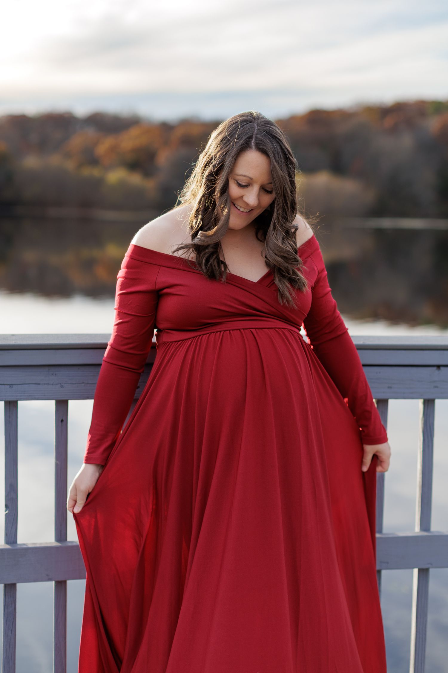 A pregnant woman in a deep red dress spreads the bottom of her dress with her hands, standing before reflective surface of Lake Fairfax.