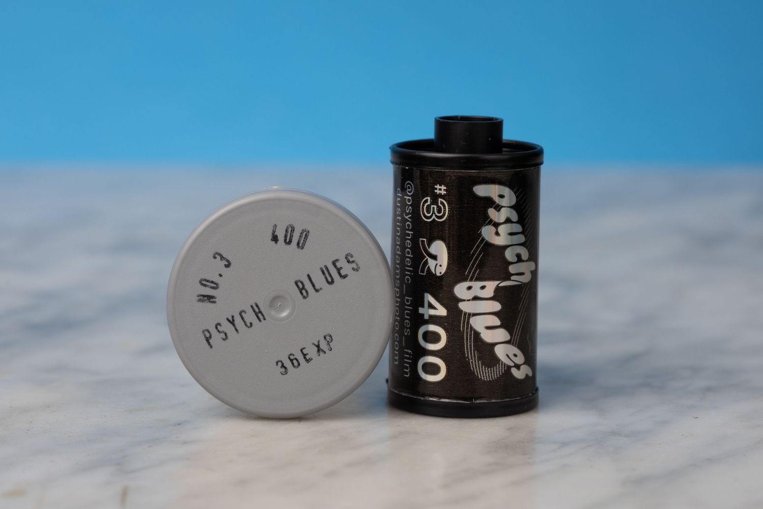 A roll of Psysch Blues 400 film sitting on a marble surface with a blue background. The plastic film canister is next to it.