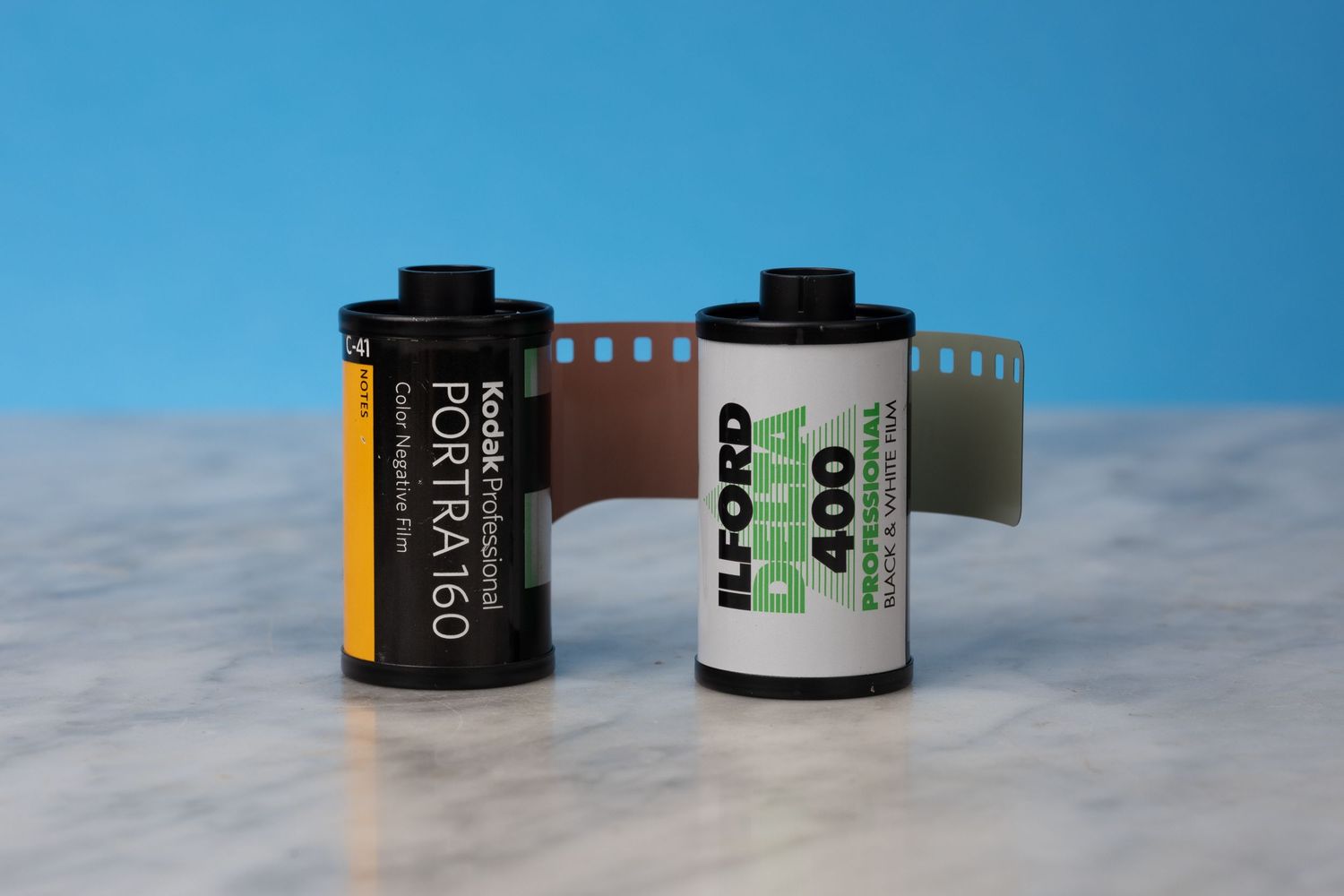 A roll of Kodak Portra 160 film and a roll of Ilford Delta 400 film sitting on a marble surface with a blue background.
