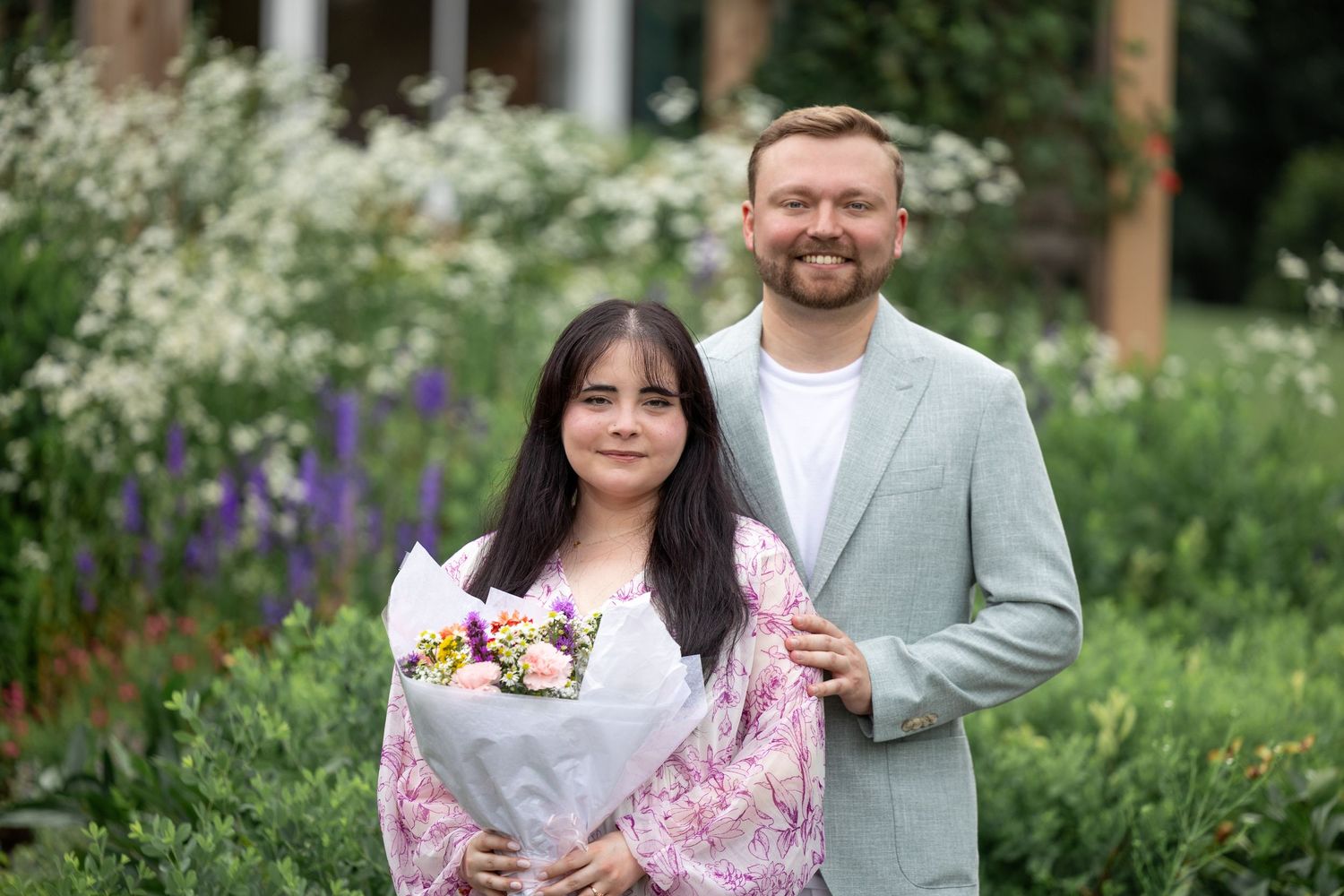 A couple poses in a green garden. The blonde man is holding the brunette woman by her shoulders.