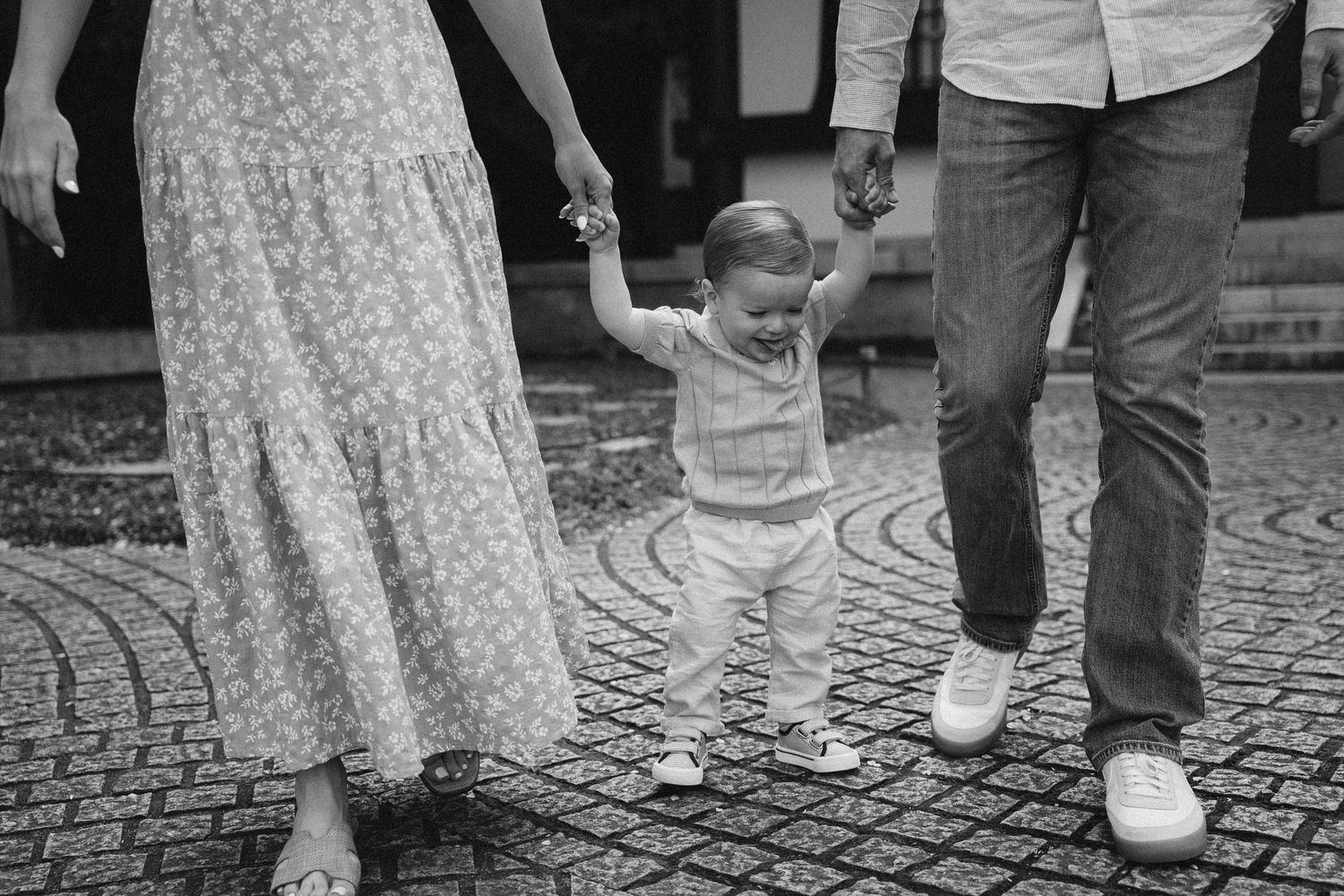 A small child takes first steps while holding hands with adults on a cobblestone street in black and white.