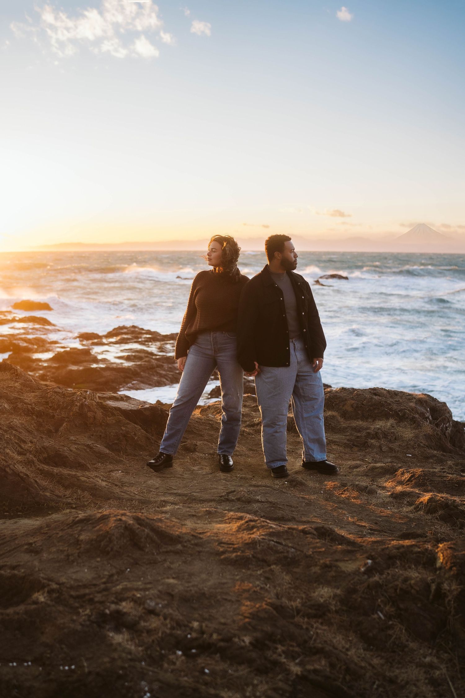 Two figures stand on rocky coastal cliffs watching the sunset over crashing ocean waves.