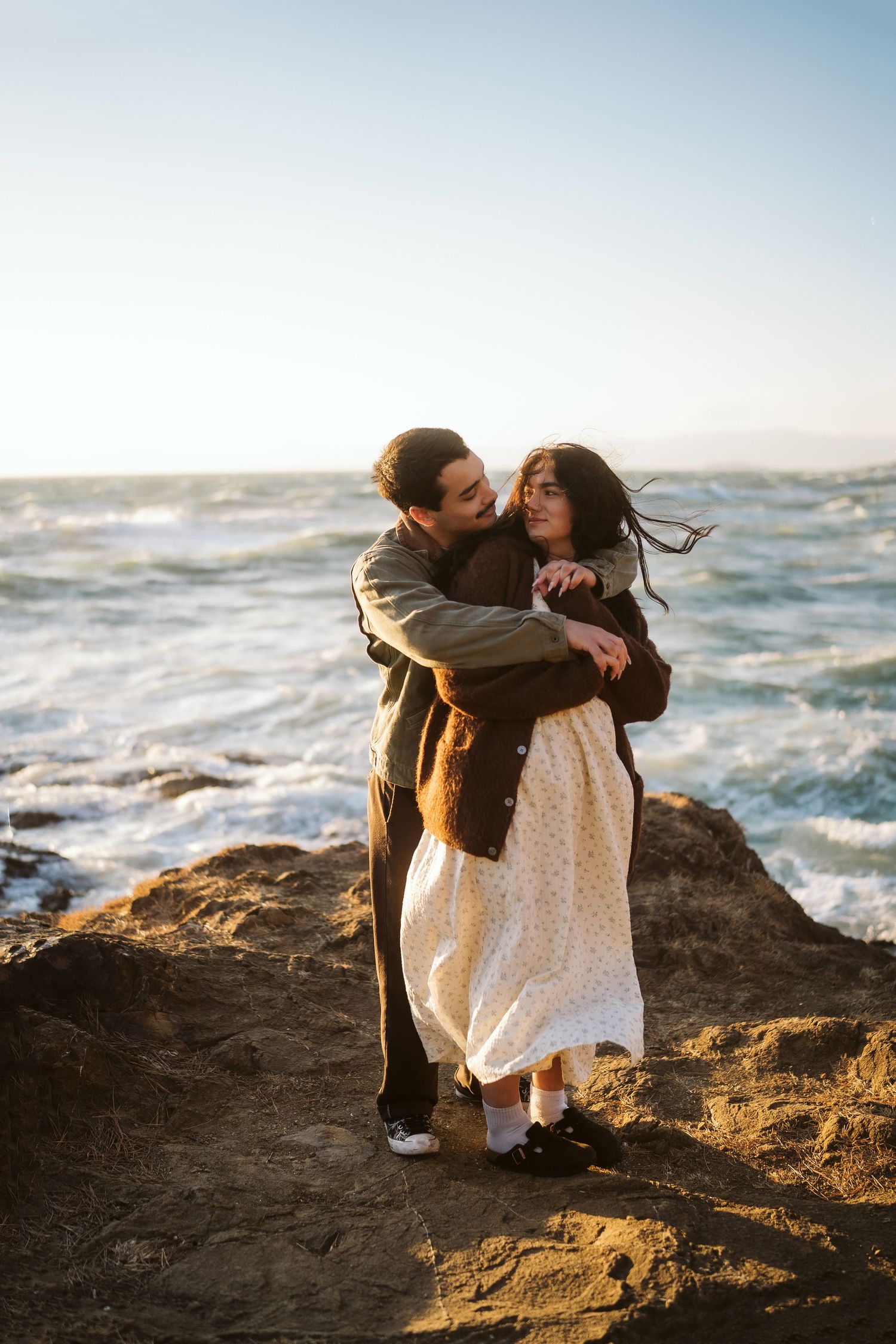 Couple embracing on rocky coastline with crashing waves and golden sunlight in the background.