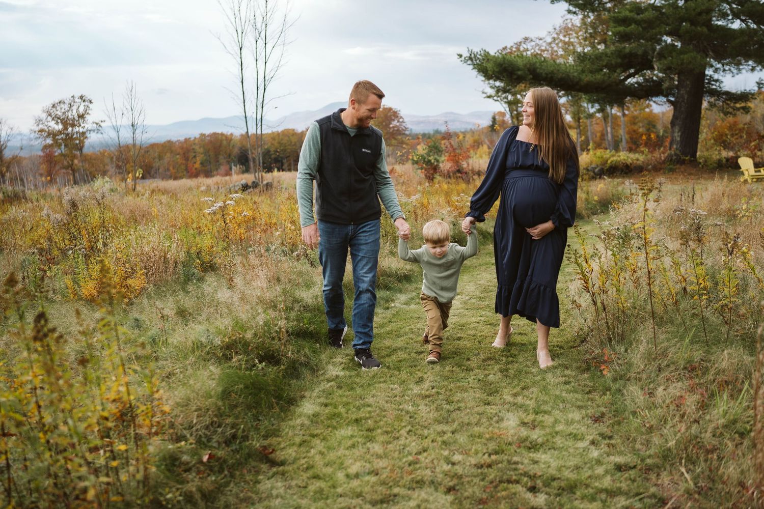Three people walking together on a grassy path through an autumn meadow surrounded by changing fall foliage.