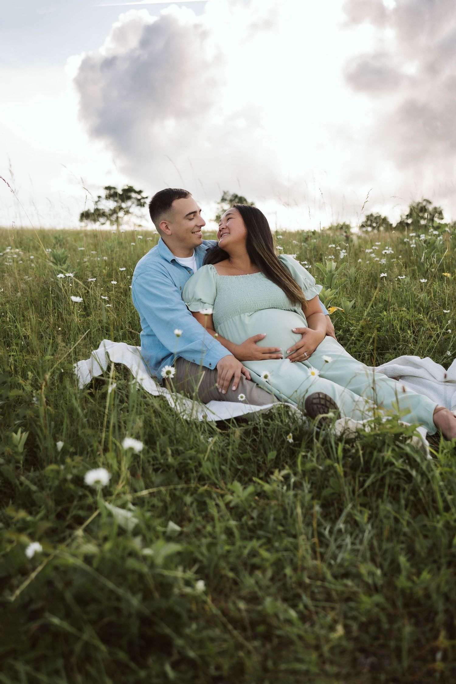 A couple sits together on a white blanket in a wildflower meadow during a cloudy summer evening.