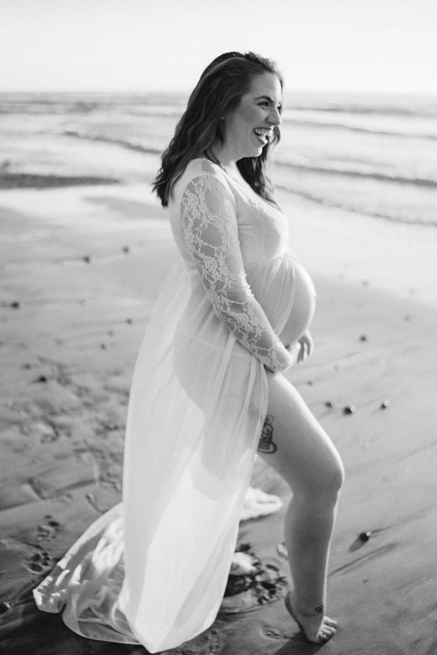 Black and white maternity photo of expecting mother in lace dress smiling on beach at sunset.