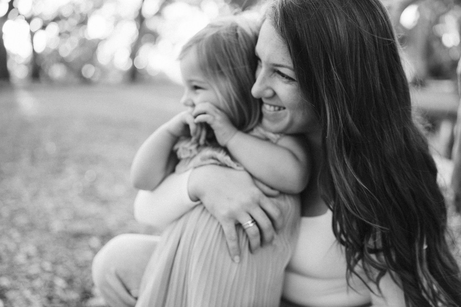 A black and white photograph shows a tender moment between a parent and child embracing outdoors on a sunny day.