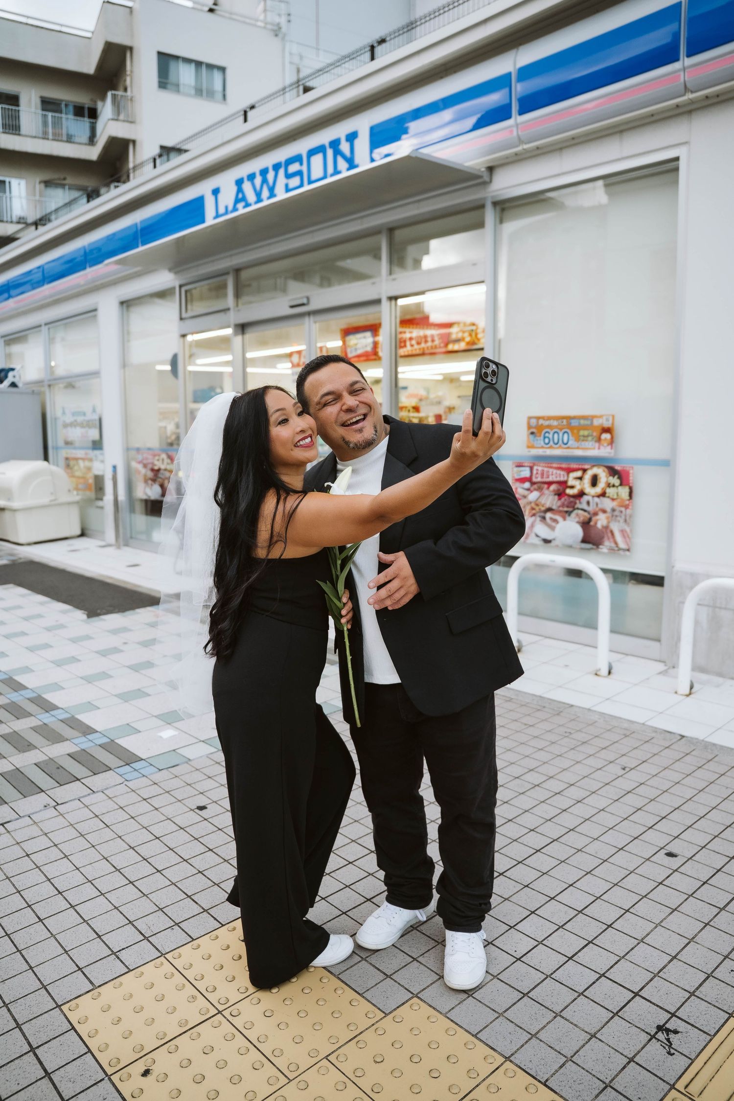 Two people in black outfits taking selfies together on a tiled floor outside a Lawson convenience store in Japan.