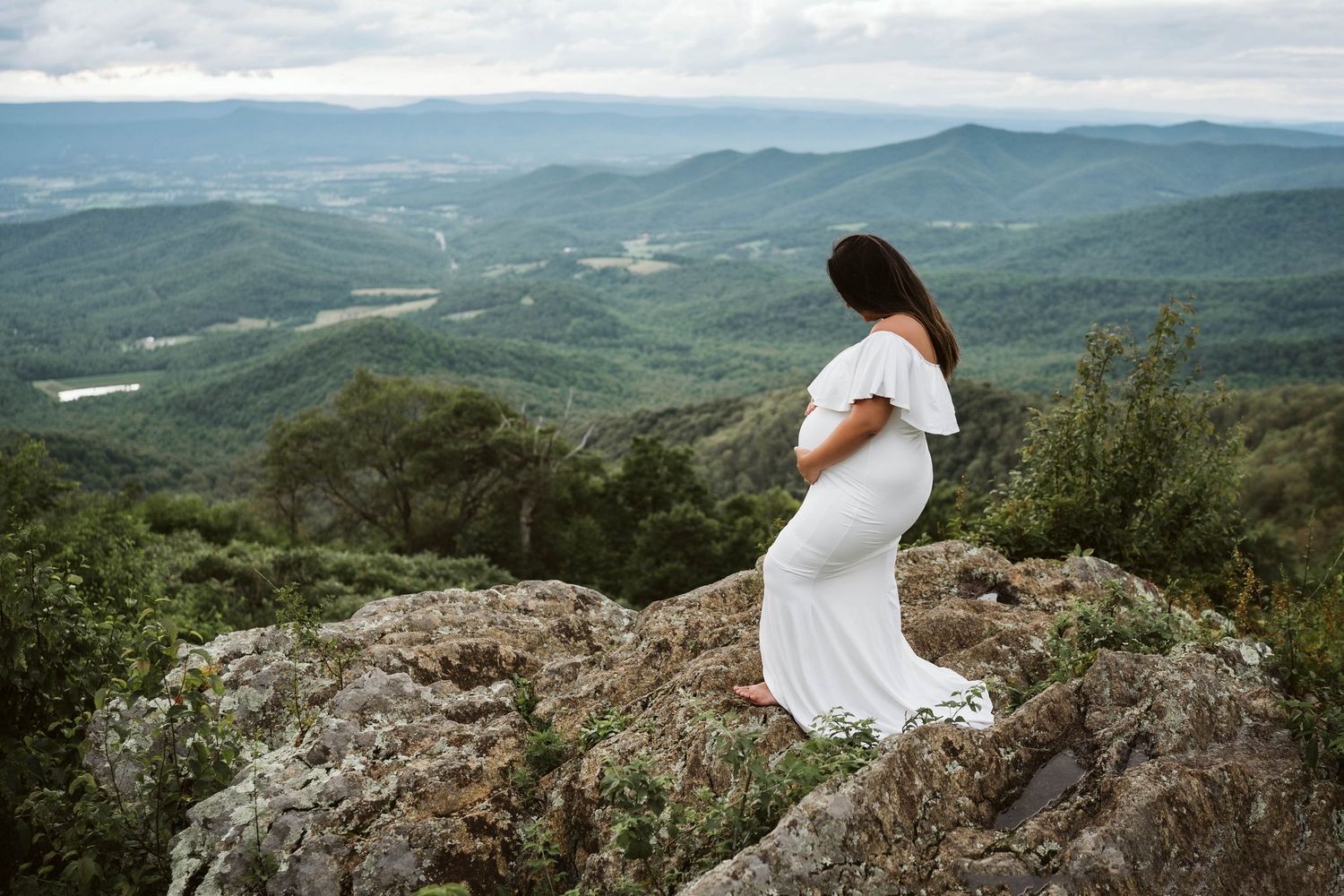 A person in a flowing white dress stands on a rocky overlook with sweeping mountain vistas in the background.