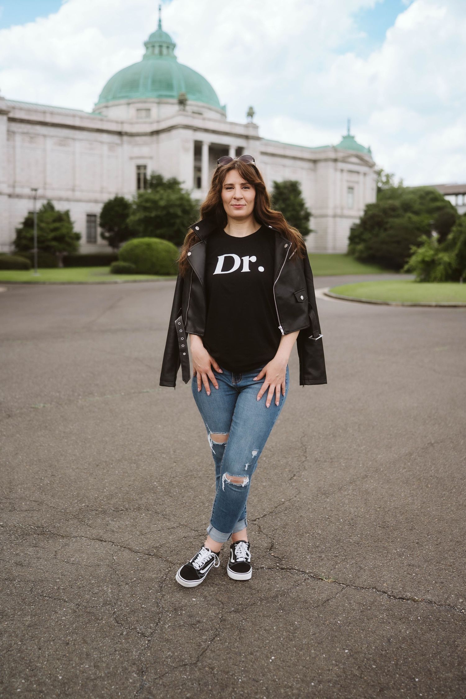 A person in casual wear poses in front of a historic building with a green dome on an overcast day.