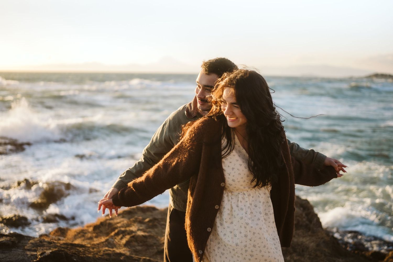A romantic sunset photoshoot along a rocky coastline with crashing waves and golden light.