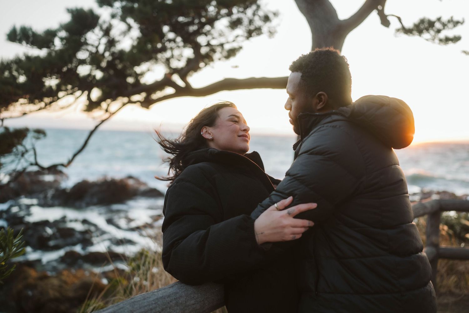A couple embraces while watching sunset over the ocean from a rocky coastal viewpoint.