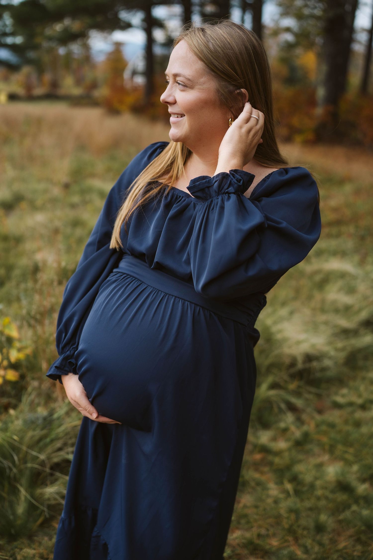 A pregnant woman wearing a navy blue dress poses outdoors during autumn with fall foliage in the background.