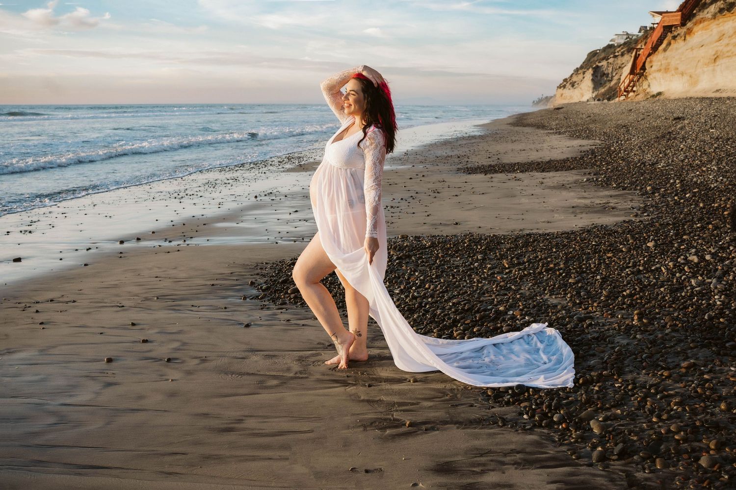 A flowing white dress billows in the ocean breeze as waves roll onto a sandy beach beneath dramatic coastal cliffs.