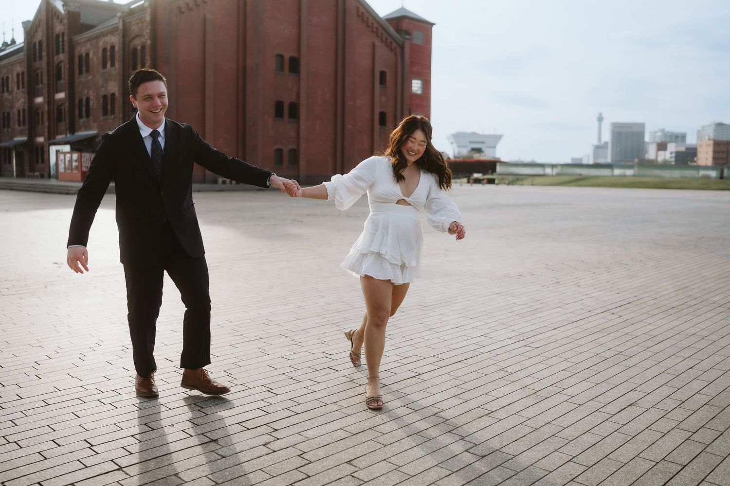 A couple in formal attire runs playfully across red brick pavement near an industrial brick building on an overcast day.