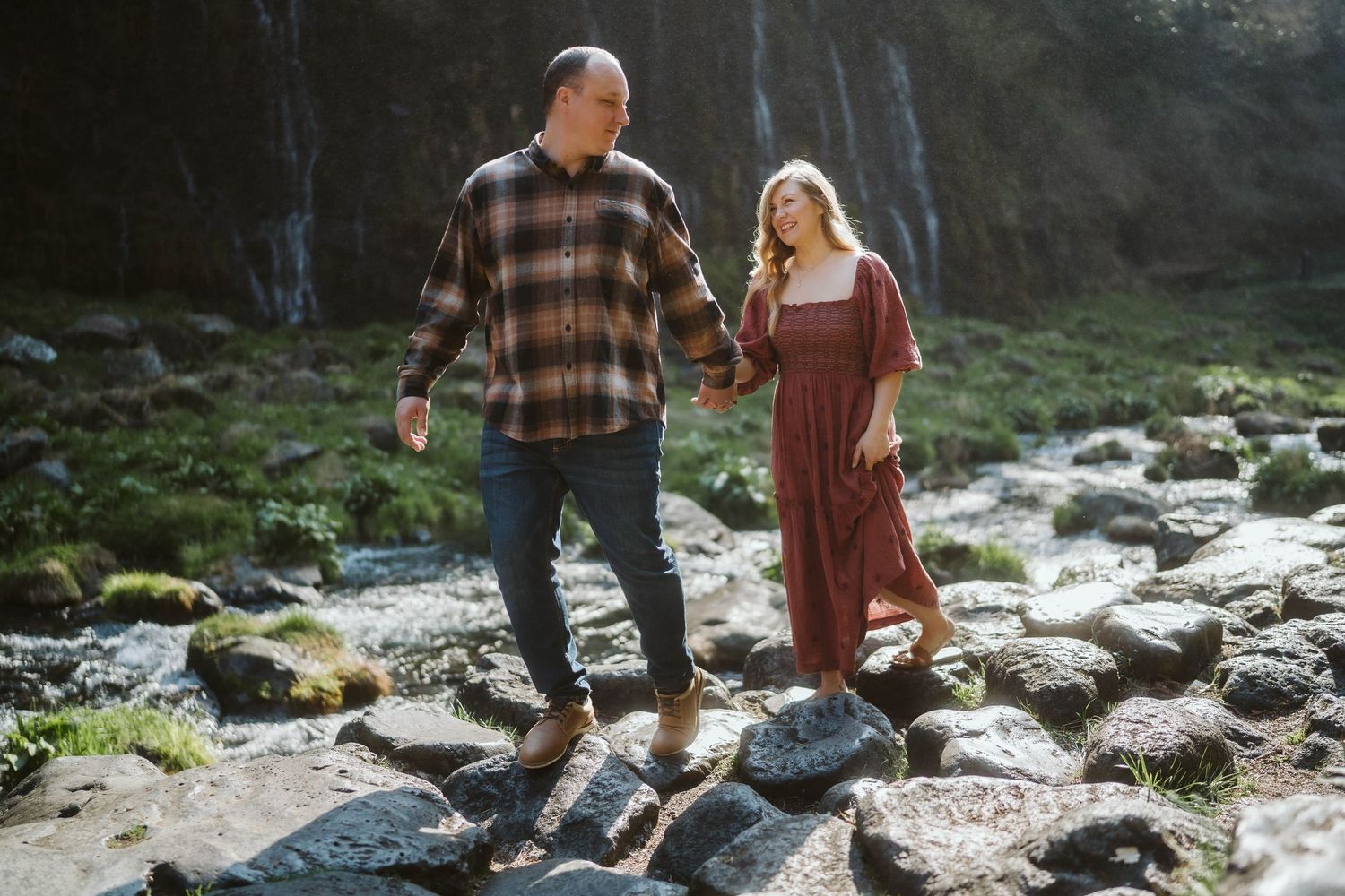 A couple walks hand in hand along a rocky stream in a scenic mountain landscape during a sunny afternoon.