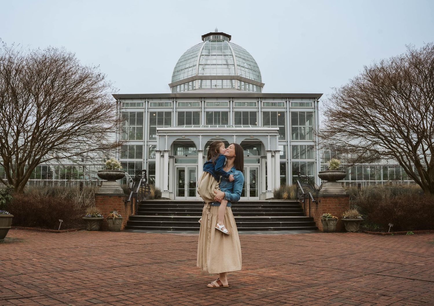 A figure stands in front of a grand glass conservatory building with a domed roof at a botanical garden on an overcast day.